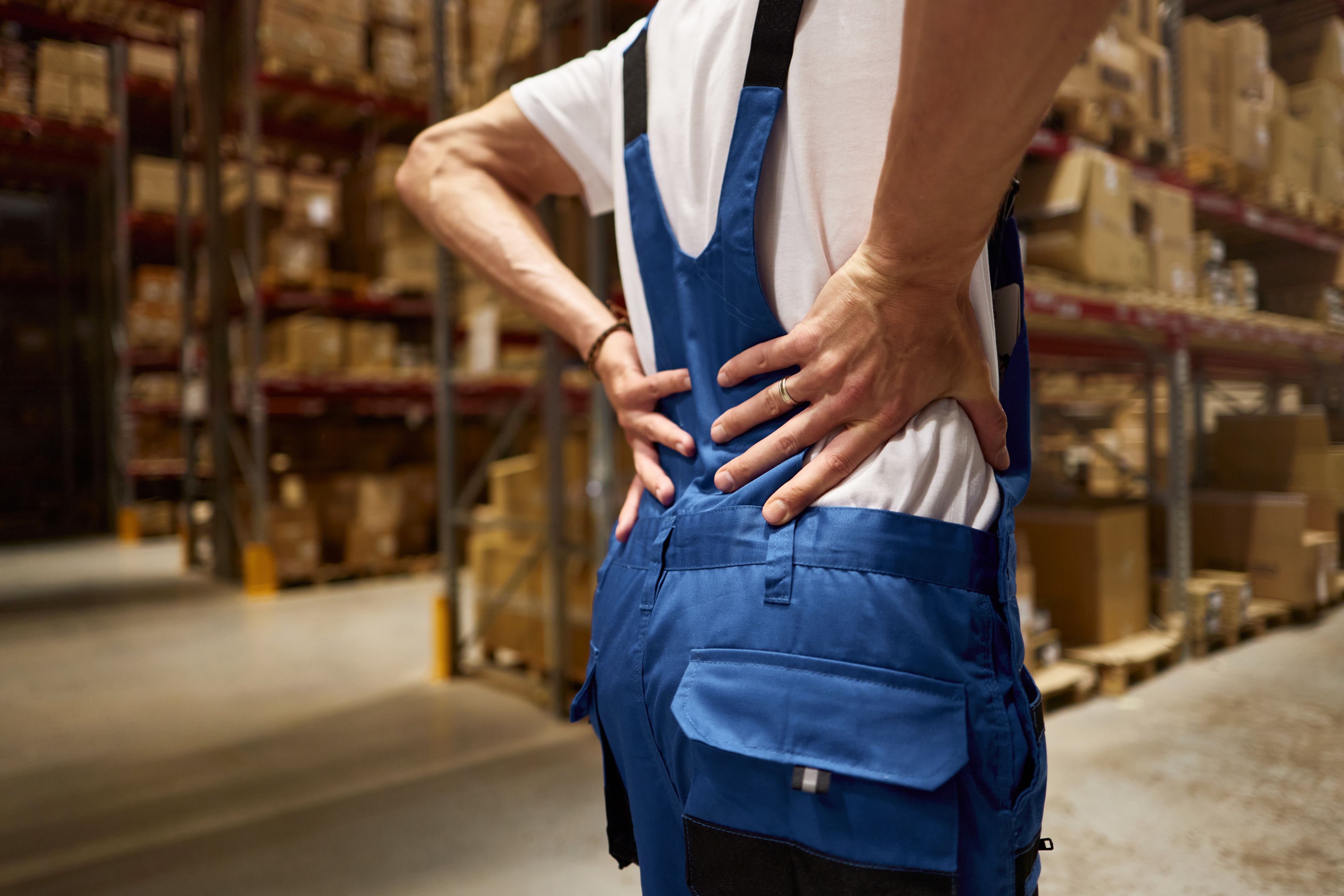 Worker in overalls holding lower back in pain at a job site, representing a workplace injury and workers' compensation issue.
