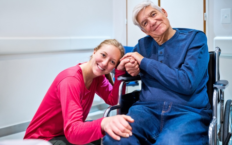 In a stock photo of grinning granddaughter beside wheelchair-bound grandfather in stock photo illustrates Attendant Care in Workers’ Compensation Cases